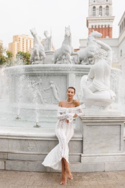 Beyaz dantel elbiseli ve incili zarif beyaz kadın Phu Quoc, Vietnam 'daki ikonik Fountain Horse' un yanında çıplak ayakla poz veriyor. Seyahat, moda veya yaşam tarzı temaları için mükemmel yüksek kaliteli görüntü.