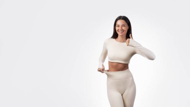 Bej renkli aktif giysili gülümseyen atletik kadın bol pantolonunu gösteriyor ve başparmağını kaldırıyor. Kavramsal fitness ve kilo kaybı portresi, sağlık, spor salonu, diyet, sağlık, promosyon ve yüksek kaliteli