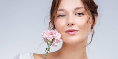 Elinde pembe karanfil çiçeği tutan gülümseyen beyaz bir kadının yakın plan portresi. Kusursuz cildi ve kendine güveni cilt bakımına, sağlığına ve kadınsı güzelliğine mükemmel bir tanıtım sağlıyor..