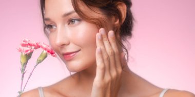 Pembe karanfili izlerken yanağına hafifçe dokunan bir kadının yakın plan resmi. Işıl ışıl ten, yumuşak tonlar ve romantik ışıklandırma bunu kozmetik ve çiçek kampanyaları için ideal kılıyor..