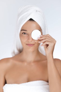 Parlak tenli genç bir kadın yüzünü temizlemek için pamuk ped kullanıyor. Havluya sarılmış halde, rahatlatıcı bir güzellik rutininden hoşlanıyor. Cilt bakımı, kaplıca ve sağlık kavramları için mükemmel..