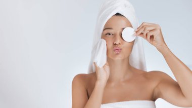 Havluya sarılmış, elinde pamuk ped ve kozmetik mendil tutarken yüzünü öpen oyuncu Asyalı kadın. Yüksek kalite güzellik, cilt bakımı, kaplıca ve kozmetik reklamcılığı ve tanıtım için sağlık portresi konsepti.