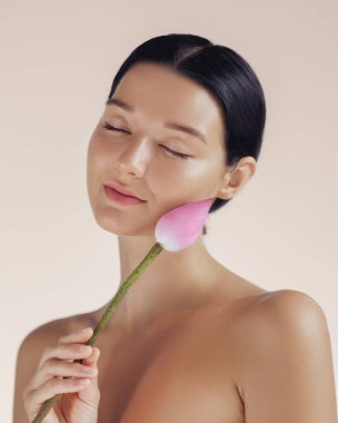 Güzel genç bir kadın yanağına pembe bir nilüfer tomurcuğu tutuyordu gözleri kapalıydı, bej bir arka planda sakince parlıyordu. Sağlık, kaplıca ve doğal kozmetik promosyonları için mükemmel..