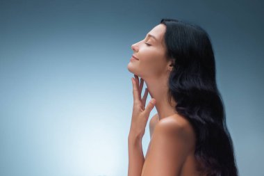 Yüzüne nazikçe dokunan pürüzsüz derili, dingin, beyaz bir kadının yan görüntüsü. Kozmetik ve sağlık reklamları için kavramsal yaşlanma karşıtı, cilt bakımı ve spa fotoğrafı.