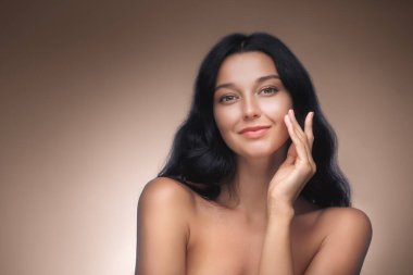 Yüzüne nazikçe dokunan, açık tenli, koyu renk saçlı, gülümseyen beyaz bir kadının yüksek kalite güzel portresi. Cilt bakımı, kozmetik reklamları ve sağlık promosyonları için ideal..