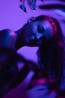 Dramatik neon ışıklarla yıkanan zarif model, başını yemyeşil tropikal yaprakların arasına eğiyor. Güzellik, gece hayatı ya da üst düzey yayın kampanyaları için ideal kavramsal moda portresi.