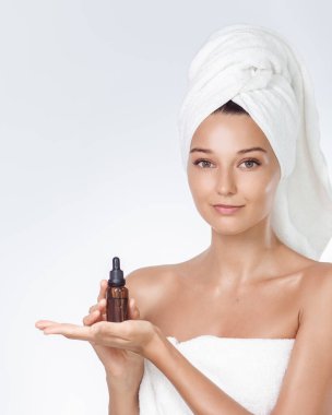 Cilt serumu sunum konsepti: Havlu türbanlı genç beyaz kadın avucunda kehribar damlatan bir şişe tutuyor, temiz beyaz stüdyo arka planı. Kozmetik reklamları, spa ve ekommerce için kaliteli bir çekim..