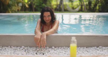 Tanned woman in orange bikini relaxing by poolside, leaning forward with a fresh juice bottle on white pebbles. Ideal for wellness, summer vacation, and healthy lifestyle themes.