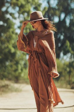 Stylish woman wearing a boho chic outfit with a wide-brimmed hat and sunglasses, walking confidently on a sunny day. Perfect for lifestyle, fashion, and summer travel concepts.
