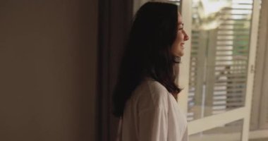 Gündelik beyaz gömlekli genç bir kadın uyanıyor, yatağından kalkıyor ve sabah güneşinde tropik bir tatil köyünün balkonuna uzanıyor. Mükemmel yaşam tarzı, tatil, sağlık ve seyahat tanıtım konsepti..