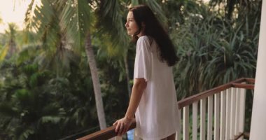 Büyük tişörtlü genç bir kadın, etrafı tropik yeşilliklerle çevrili bir balkondan neşeyle dışarı bakar. Eğlenceli ve neşeli bir an, yumuşak sabah ışığında yakalanır. Seyahat reklamları için mükemmel..