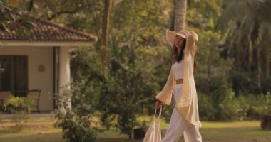 Dalgalı bohem tarzı giyinmiş ve hasır şapkalı bir kadın güneşle yıkanmış sakin bir tatil köyü bahçesinde yürüyor. Seyahat, lüks tatil köyü deneyimleri ve bohem modası için mükemmel..