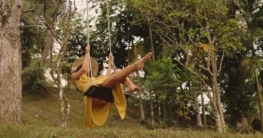 Dalgalı sarı elbiseli neşeli kadın tropikal yeşilliklerin arasında sallanıyor. Seyahat, moda ya da sağlık reklamları için çekilen güzel, kaygısız bir yaşam tarzı anı. Yüksek kaliteli video.