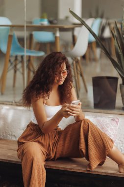 Şık bir kafe terasında otururken cep telefonuyla mesajlaşan mutlu genç bir kadın. Modern kentsel yaşam ve reklam için iletişim, sosyal yaşam tarzı ve dijital bağlantı kavramı.