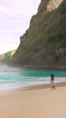 Bali 'deki Kelingking Sahili' nin tropikal kıyılarında sabah ışığında yürüyen zarif bir kadın. Seyahat reklamları, özgürlük temaları veya moda kampanyaları için ideal kavramsal dikey görüntüler.