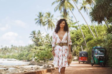 Sri Lanka Hiriketiya sahil yolcusu, palmiyeler ve renkli tuk kuşlarıyla kaplı güneşli bir sahil yolunda yürüyor, okyanus esintisi arkamızda. Turizm reklamları, seyahat blogları, sosyal medya, hedef kampanyalar için ideal.