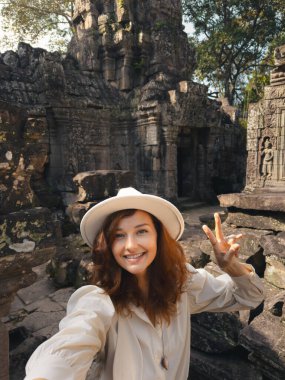 Kamboçya 'nın Angkor Wat kentindeki Ta Som tapınağında selfie çekerken gülümseyen ve barış işareti yapan beyaz bir kadın. Seyahat blogları, turizm reklamları ve tatil tanıtımları için kavramsal imaj.