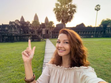 Genç bir kadın Angkor Wat tapınağını işaret ederken sabahın erken saatlerinde selfie çekiyor. Yüksek kaliteli stok imajı seyahat blogları, turizm reklamları ve kültürel keşif kampanyaları için mükemmeldir..