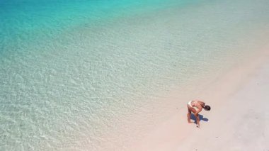 Kafkasyalı bir kadın tropikal kumlu plajda güneş kremi sürüyor, temiz turkuaz suyla üst hava manzaralı. Güneş koruma kavramı, cilt bakımı, yaz tatili ve sağlıklı yaşam tarzı..