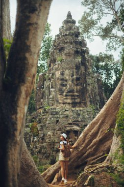 Bayon Face Kulesi kadın yolcusu Angkor Thom, Siem Reap, Kamboçya 'da dev ağaç kökleri altında duruyor ve Khmer kapısına bakıyor. Reklamlar, bloglar ve başyazılar için kavramsal ve yüksek kaliteli turizm imajı.
