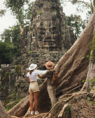 Bayon, Angkor, Kamboçya 'da haritalı iki gezgin, yayılan köklerin arasında oyulmuş taş bir yüzü inceler. Turizm pazarlaması, kültür yazıları, bloglar ve macera reklamları için yüksek kalite, kavramsal imaj.