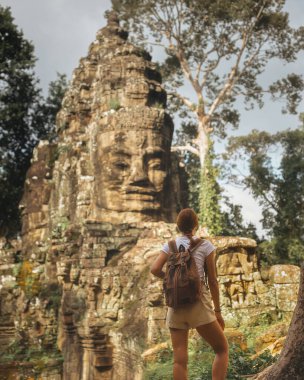 İnce Kafkas kadın maceracı, bir ağacın köklerinde duruyor ve Kamboçya 'daki ünlü Bayon Tapınağı kapısına bakıyor. Seyahat için kavramsal fotoğraf, orman gezisi ve kayıp miras..