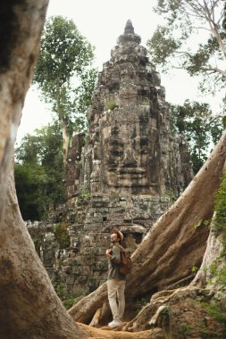 Şapka ve sırt çantası takan genç kaşif Angkor Thom, Kamboçya 'daki antik Bayon yüz tapınağının önünde dev kökler arasında duruyor. Çevre rehberleri, kültür blogları, turları ve
