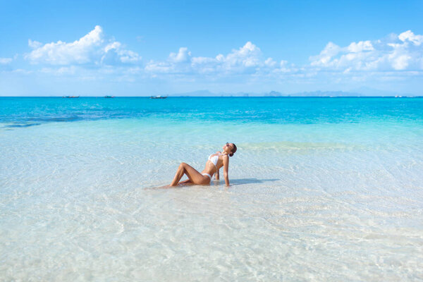 Young bikini model lounges in shallow, crystal clear ocean water under a sunny sky. Great for vacation, travel promo, beach lifestyle, summer fashion, and sensual branding.