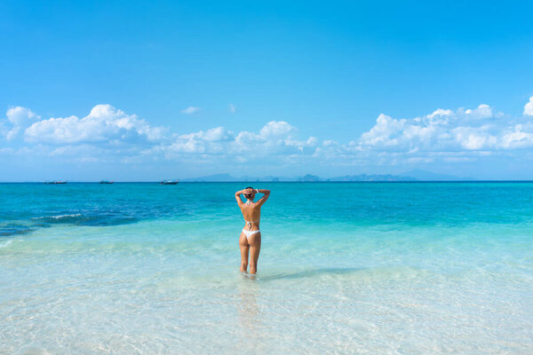Rear view young woman in white bikini stands in shallow turquoise sea on a Thai island beach, arms raised behind head. Concept: lifestyle travel, fashion, wellness, high quality ad for tourism promo.