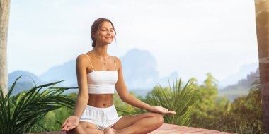 Yumuşak dağ manzaralı sakin bir tropikal bölgede yoga meditasyonu yapan bir kadın, sağlıklı markalar, yaşam tarzı görselleri, seyahat reklamları ve meditasyon için mükemmel kavramsal yüksek kaliteli bir tanıtım fotoğrafı.