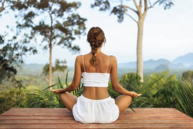 Genç beyaz kadın ahşap bir güvertede nilüfer pozisyonunda oturuyor, yemyeşil orman ve uzak dağ zeminiyle meditasyon yapıyor. Sağlık, farkındalık, kaplıca ve geri çekilme reklamları için ideal..
