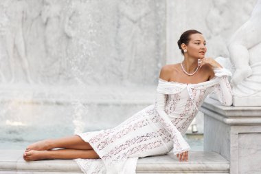 İncili, beyaz dantel bir elbise giymiş zarif bir kadın Phu Quoc 'taki at heykellerinin yer aldığı büyük bir çeşmenin mermer çıkıntısına yalınayak uzanır. Yüksek kalite seyahat, güzellik ya da gelin editörlüğü imajı.