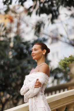 Dantel bir gelinlik içinde zarif bir gelin, yapraklı bir ortamda, yumuşak ve özgüvenli bir şekilde gülümsüyor. Moda, gelin pazarlaması, aşk, yaşam tarzı ve romantik görsel temalar için ideal.