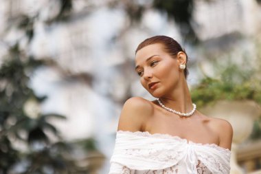 Omuzları açık beyaz dantelli kadın portresi, yumuşak bokeh yeşilliğine karşı sakin bir zarafetle bakıyor. Gelin yazıları, güzellik reklamları, lüks markalar için ideal..