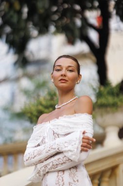İnci kolyeli ve küpeli beyaz dantelli bir kadın lüks otel merdivenlerinde çapraz kollarıyla poz veriyor ve yumuşak bokeh yeşiliyle poz veriyor. Gelin modası reklamları için ideal, zarif markalaşma.