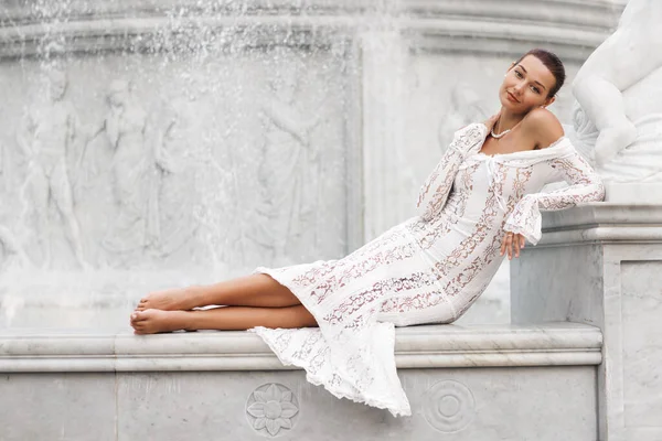 Beyaz dantel elbiseli zarif bir kadın mermer çeşmede poz veriyor, lüksü, romantizmi ve zamansız güzelliği ifade ediyor, gelin moda yazıları, yaşam tarzı blogları, seyahat reklamları ve yaratıcı markalaşma için ideal..