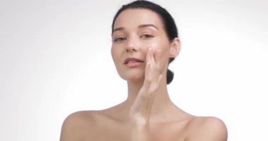 Güzel, beyaz bir kadın beyaz bir stüdyoda parıldayan, nemli bir ten göstererek yanaklarına hafifçe dokunuyor. Kozmetik ürün reklamcılığı için kaliteli cilt bakımı, sağlık ve güzellik konsepti.