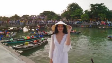 Beyaz elbiseli, konik Vietnamlı şapkalı, çekici beyaz kadın canlı Hoi An nehrinin kıyısında tekneler ve kalabalıklarla dururken sıcacık gülümsüyor. Seyahat, yaşam tarzı ve tanıtım kullanımı için ideal.