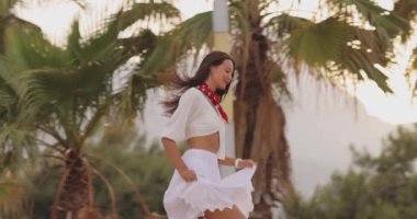 Neşeli kadın, tropikal bir tatil köyü bahçesinde ağır çekimde koşuyor. Eteğin esintide dalgalanması gibi gülümsüyor. Turizm reklamları, moda videoları ve tasasız tatil hikayeleri için mükemmel..