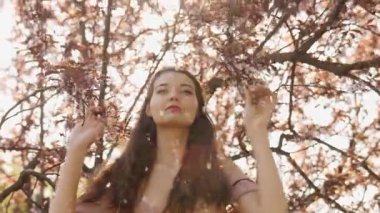 Beyaz kadın sakura ağaçlarının altında zarifçe poz veriyor. Etrafına yapraklar dökülüyor. Güzellik, moda, ilkbahar ya da doğa temalı kampanyalar için ideal yavaş çekim sahnesi.