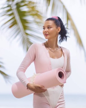 Güneşli plajda elinde pembe bir yoga minderi tutan genç bir kadın. Doğal ışık yaşam tarzı portresi, sakin sağlık durumu, fitness reklamları, inzivaya çekilmeler, bloglar ve sosyal medya için ideal.
