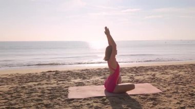 Pembe aktif giysili zayıf kadın sabah güneşinde okyanus kıyısında plaj minderinde yoga meditasyonu yapıyordu. Fitness, geri çekilme ya da yaşam tarzı reklamları için kavramsal sağlık ve farkındalık imajı.
