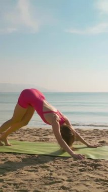 Drone, Vietnam 'da Da Nang' da güneşli bir plajda yoga yapan pembe spor giysili genç bir kadının etrafında dönüyor. Sağlık, seyahat veya fitness ile ilgili medya projeleri için huzurlu bir manzara.