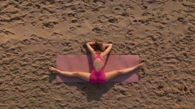 Pembe kıyafetli atletik bir kadının kumsalda yoga minderinde bacak aralarını açarken drone görüntüsü. Fitness, sağlık, inziva ve açık hava sağlık promosyonları için kavramsal fotoğraf.