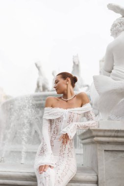 Omuz üstü beyaz dantelli gelinlik içindeki muhteşem gelin heykel detaylarıyla süslü bir mermer çeşmenin yanında poz veriyor. Moda, gelin yazıları, lüks düğün ve romantizm kavramları için ideal.