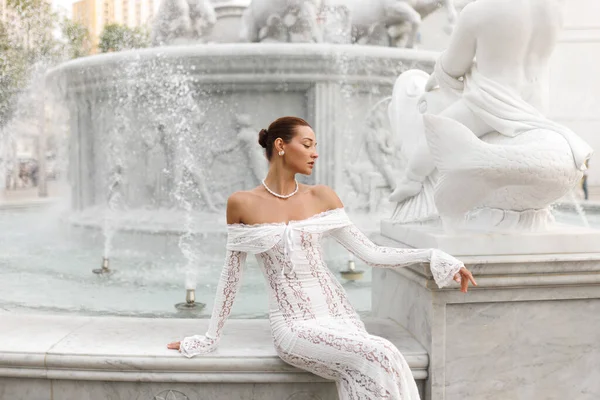 Omuz danteli elbisesiyle genç, zarif bir gelin klasik heykelle mermer bir çeşmenin yanında poz veriyor. Lüks düğünler, romantizm, zarafet ve gelin yazıları için kavramsal.