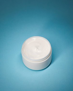 Mavi arka planda beyaz kozmetik nemlendiricili açık yüz kremi kavanozu, temiz stüdyo ışıkları ve fotokopi alanı olan yumuşak gölgeler. Cilt bakımı reklamları, ambalaj modelleri, ekommerce ve web pankartları için ideal.