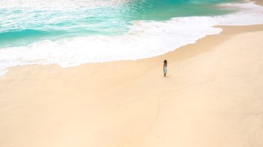 Yumuşak turkuaz dalgalarla sakin kumlu bir plajda tek başına yürüyen bir kadın, seyahat, sağlık ve yaz temaları için ideal, bloglar, reklamlar, web siteleri ve sosyal medya için mükemmel kaliteli görüntü.