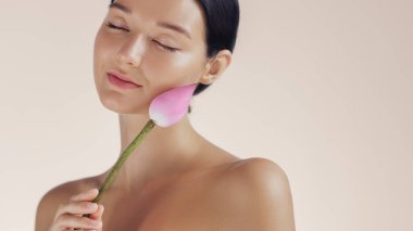 Gözleri kapalı zarif bir kadın yüzünde pembe bir nilüfer tomurcuğu tutuyor ve doğal güzelliği ve sağlığı vurguluyor. Spa, cilt bakımı, güzellik ve lüks kozmetik için mükemmel.
