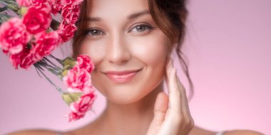 Canlı pembe karanfillerden oluşan bir buket tutarken yumuşak bir şekilde gülümseyen, ışıl ışıl cildi ve doğal makyajı olan neşeli genç bir kadının yakın plan portresi. Yumuşak pastel pembe arka plan sıcaklık katar.